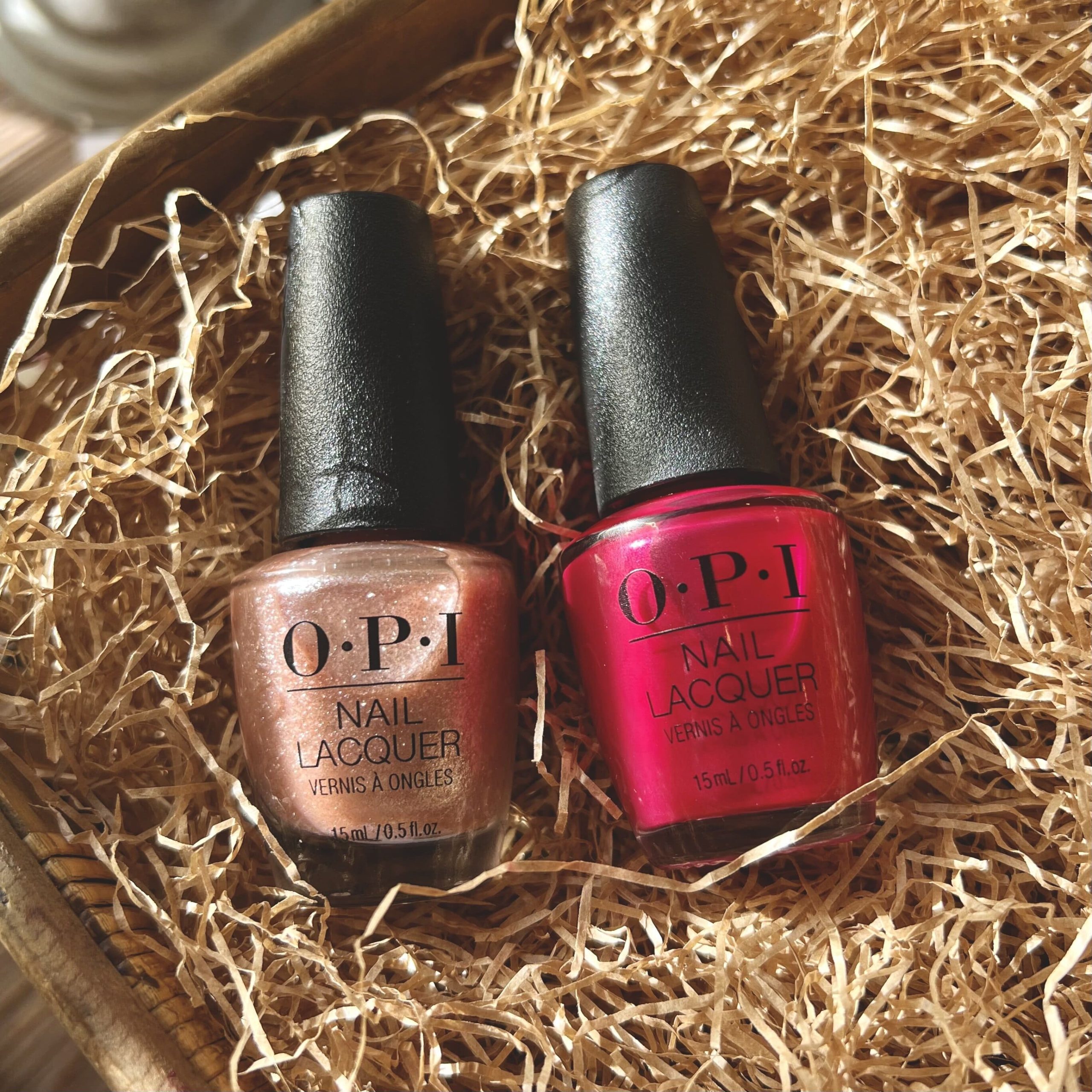 Two OPI nail polishes in a shimmery red and a metallic rose gold shade sit in a woven basket full of brown crinkle paper. 