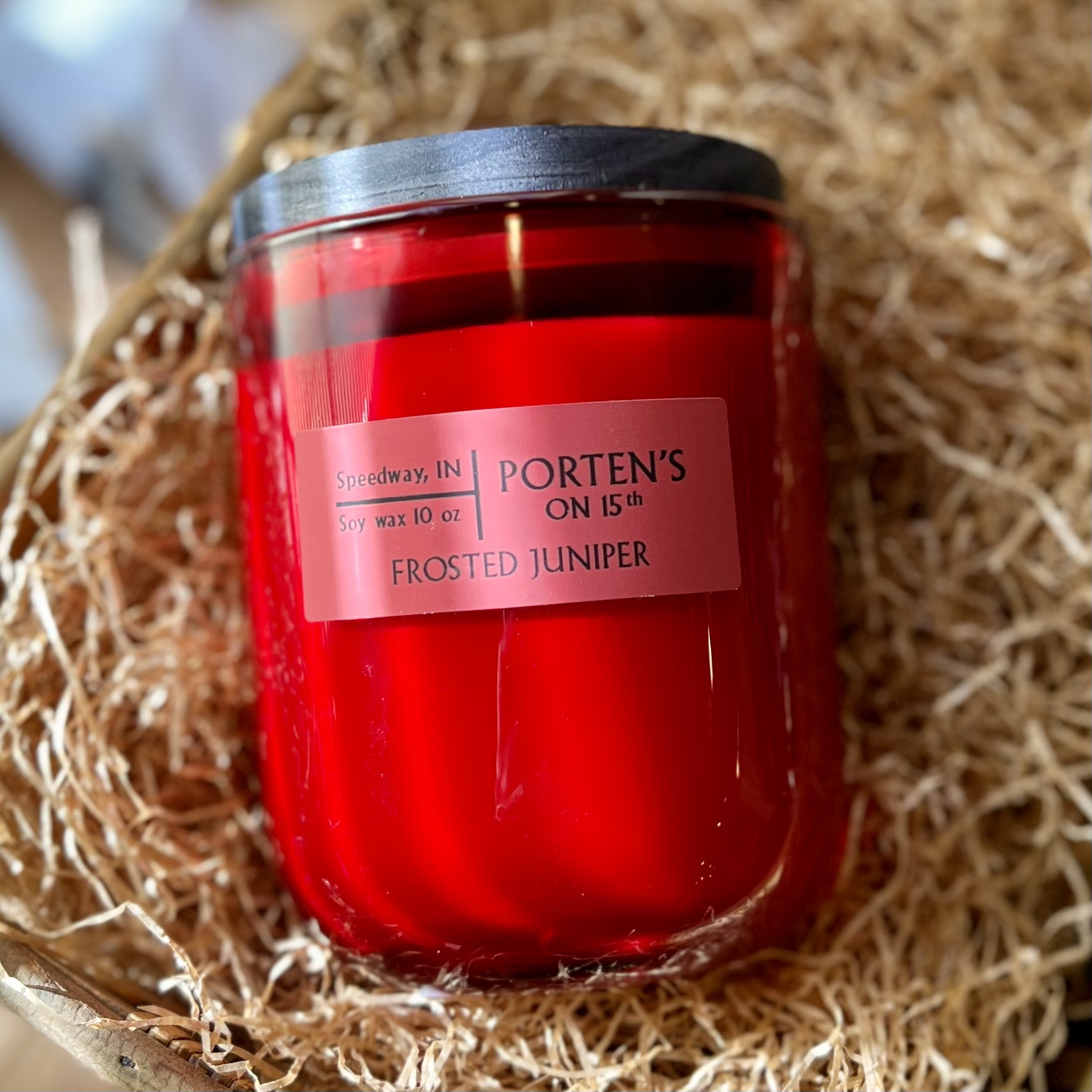 Porten's on 15th Candle Company's soy wax candle in the scent Frosted Juniper sits in a woven basket full of brown crinkle paper.