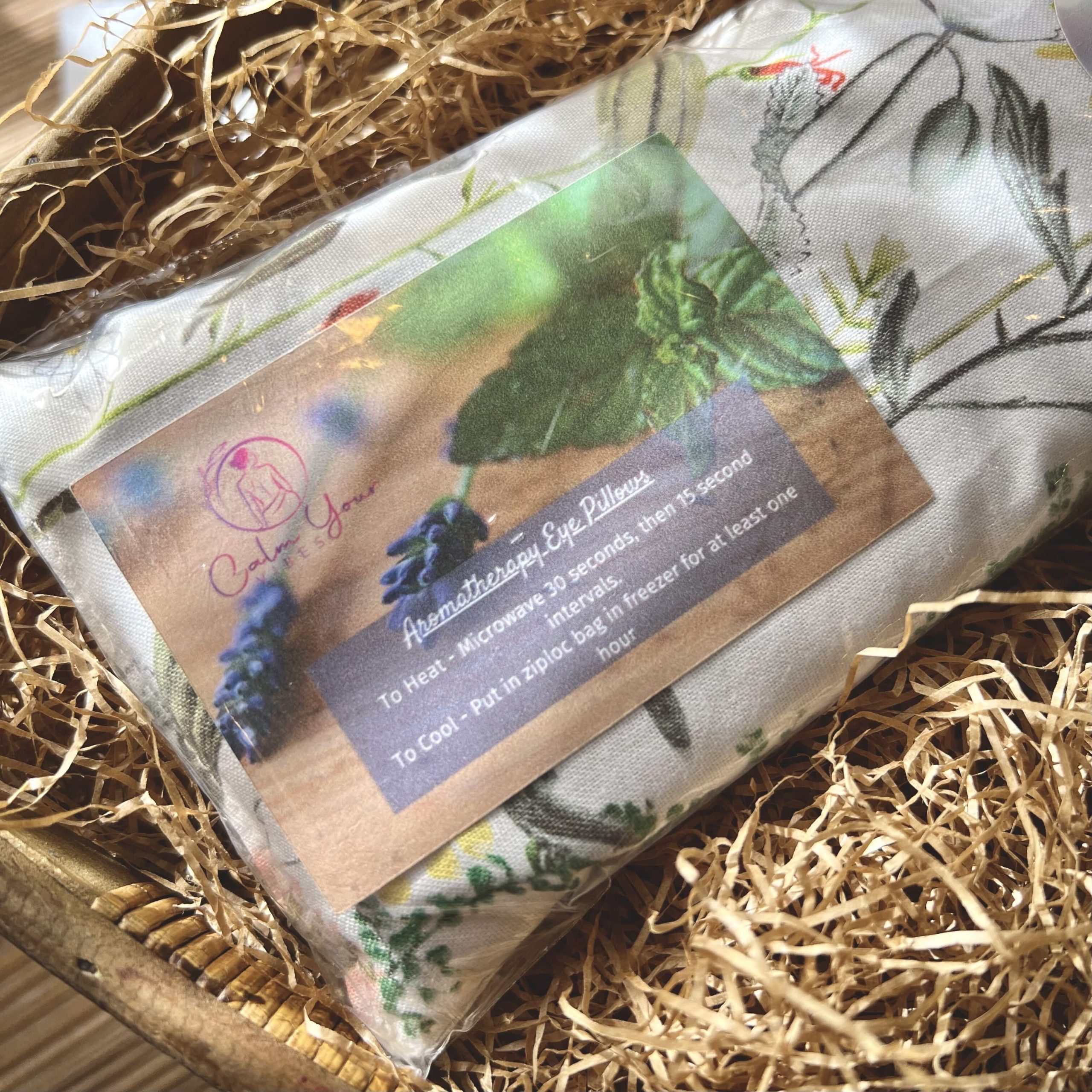 Calm Your Vibe's aromatherapy weighted eye pillow in the scent lavender sits in a woven basket full of brown crinkle paper. 