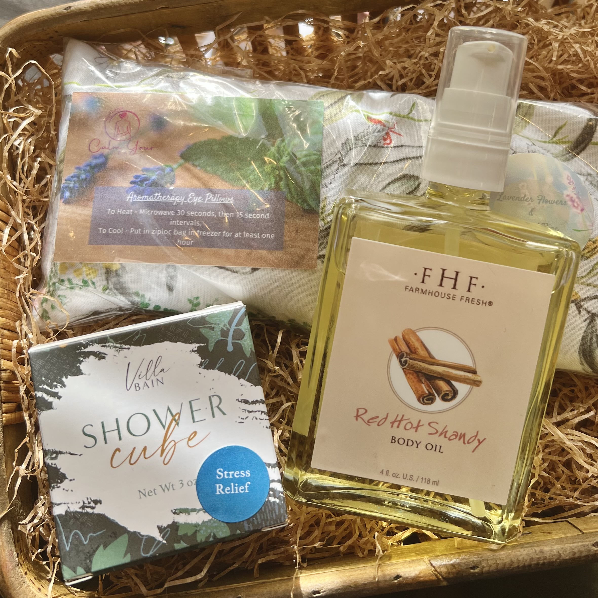 FarmHouse Fresh Red Hot Shandy body oil, Villa Bain stress relief shower cube, and Calm Your Vibe lavender aromatherapy weighted eye pillow sit in a woven basket full of brown crinkle paper.