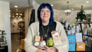 Becca, our guest services manager with blue hair and glasses stands inside Tyler Mason Salon Spa, holding various beauty and skincare products, with shelves and products visible in the background. - Tyler Mason Salon + Spa in Indianapolis, IN