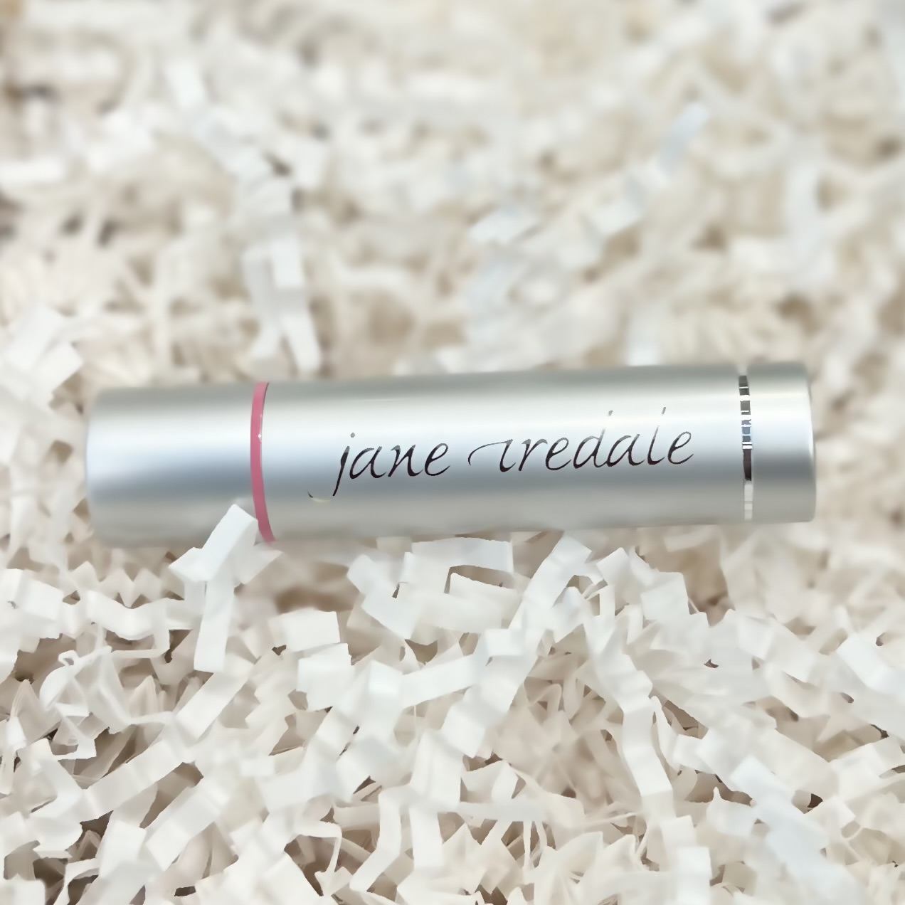 Jane Iredale's Lip Drink lip balm in the shade flirt sits in a basket of white crinkle paper.