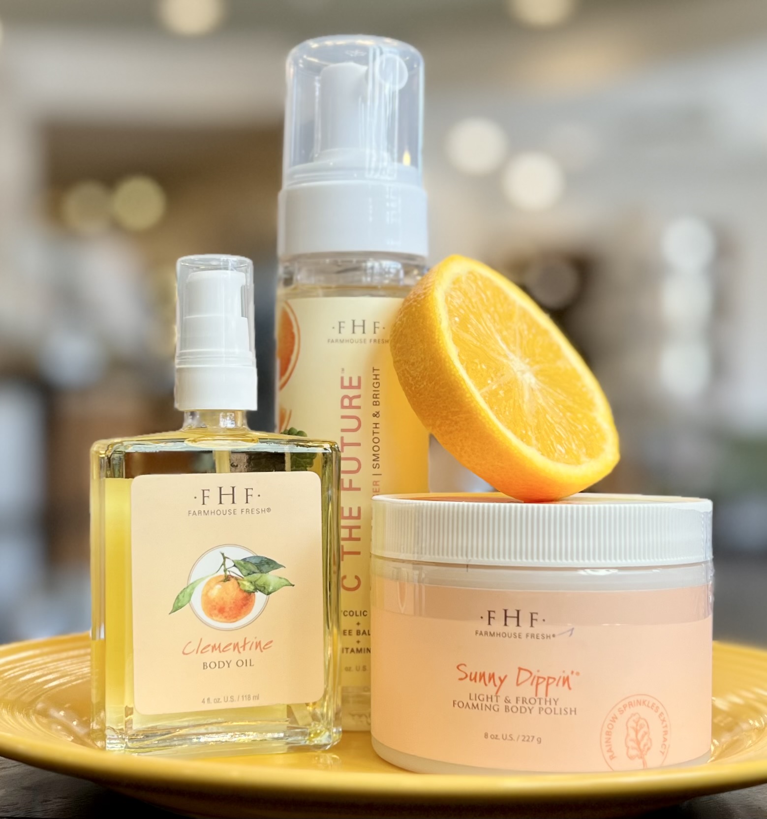 FarmHouse Fresh's Clementine Body Oil, Sunny Dippin' Foaming Body Scrub, and C the Future Facial Cleanser sit on a yellow plate with an orange slice.