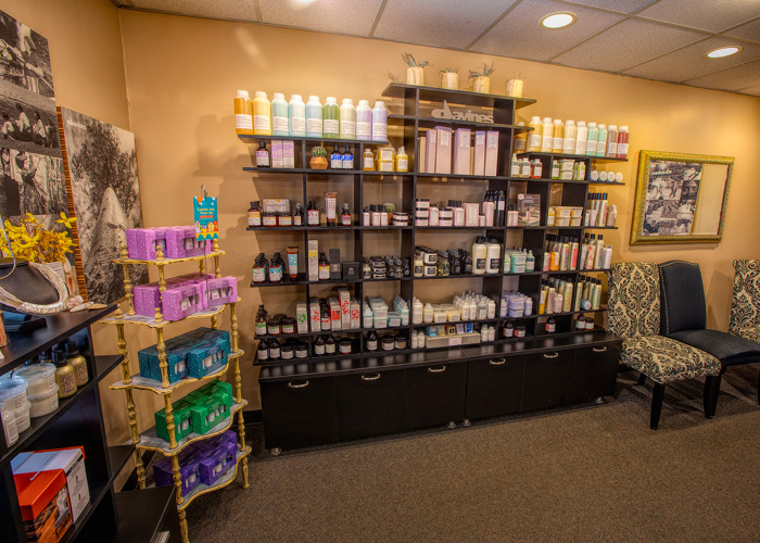 Shelves filled with various hair and beauty products are displayed against a wall in a salon, with two patterned chairs and framed photos nearby. - Tyler Mason Salon + Spa in Indianapolis, IN