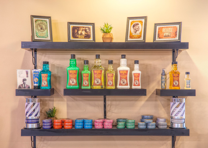 Three wall shelves display framed photos, small plants, bottles of Reuzel grooming products, and stacks of colorful hair pomade tins. - Tyler Mason Salon + Spa in Indianapolis, IN