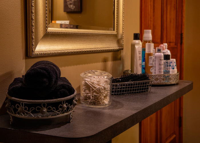 A bathroom countertop with rolled black towels, a jar of cotton swabs, hairbrushes, and various toiletries arranged in trays, beneath a framed mirror. - Tyler Mason Salon + Spa in Indianapolis, IN