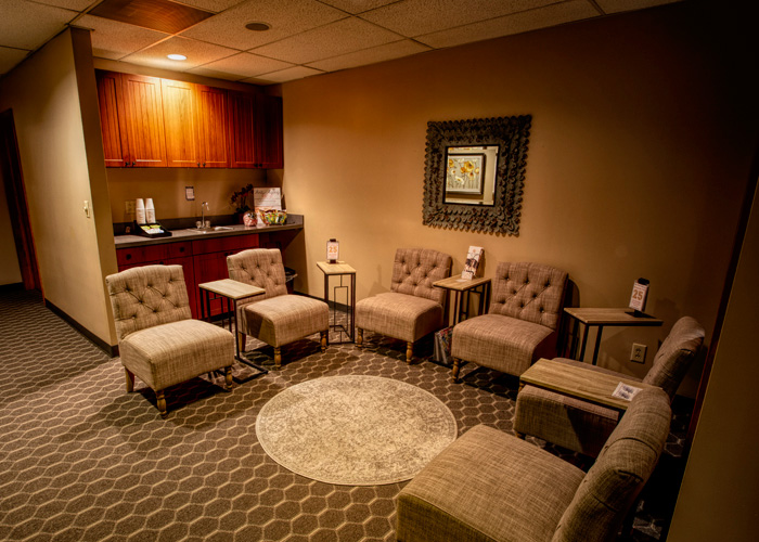 A small waiting room with eight upholstered chairs, side tables, a circular rug, a wall mirror, refreshments on a wooden counter, and warm ambient lighting. - Tyler Mason Salon + Spa in Indianapolis, IN