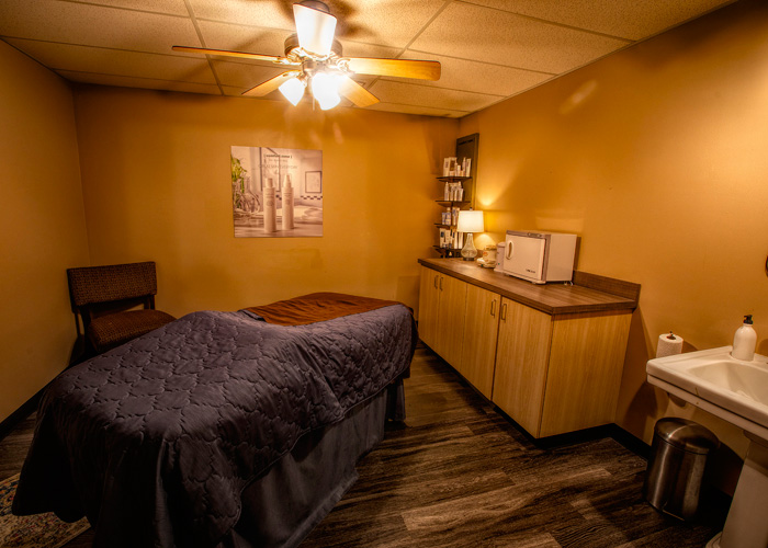 A massage therapy room with a bed, brown walls, overhead ceiling fan, cabinets, a sink, and a chair in the corner, softly lit with warm lighting. - Tyler Mason Salon + Spa in Indianapolis, IN