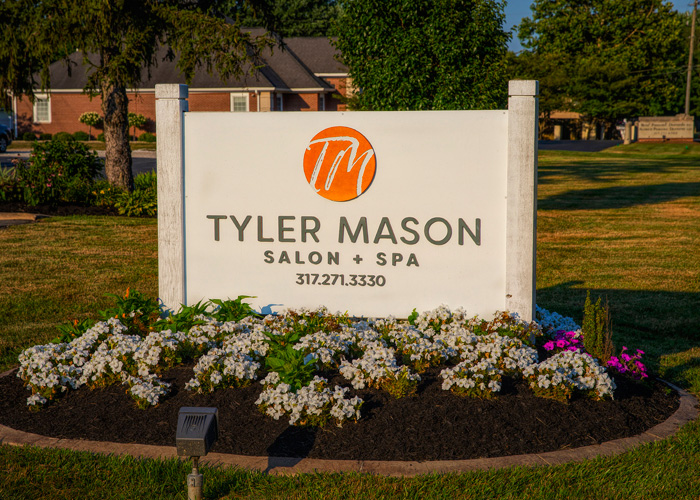 A white sign for Tyler Mason Salon + Spa with orange logo and contact number, surrounded by flowers and grass, with a building and trees in the background. - Tyler Mason Salon + Spa in Indianapolis, IN