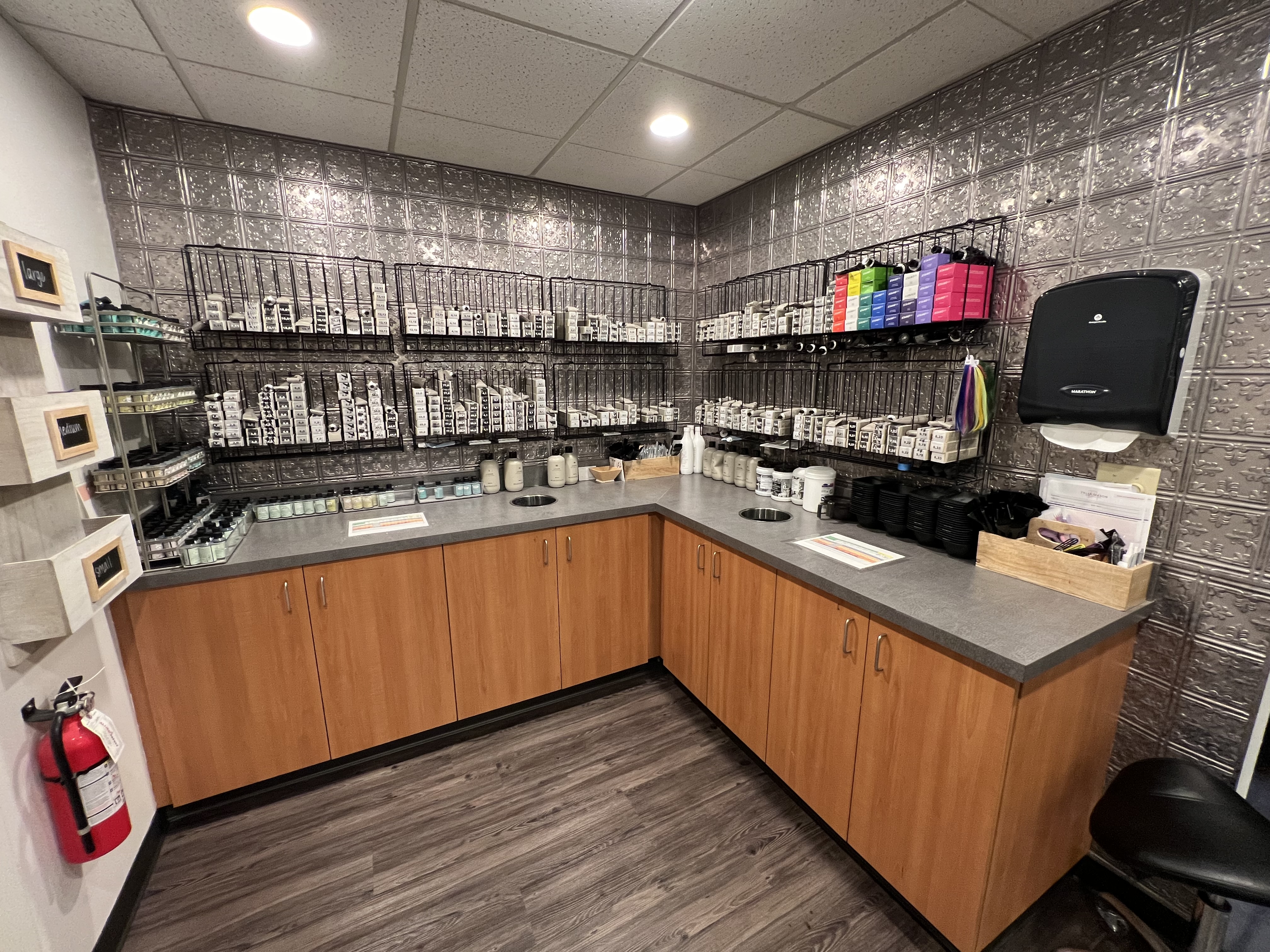 A well-organized room with wooden cabinets, gray counters, labeled containers on wire racks, a wall-mounted paper towel dispenser, and a fire extinguisher. - Tyler Mason Salon + Spa in Indianapolis, IN