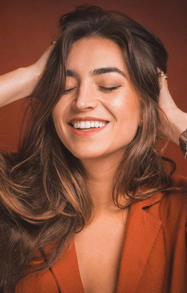 Woman with long brown hair smiles with eyes closed, hands in her hair, wearing an orange jacket against a brown background. - Tyler Mason Salon + Spa in Indianapolis, IN
