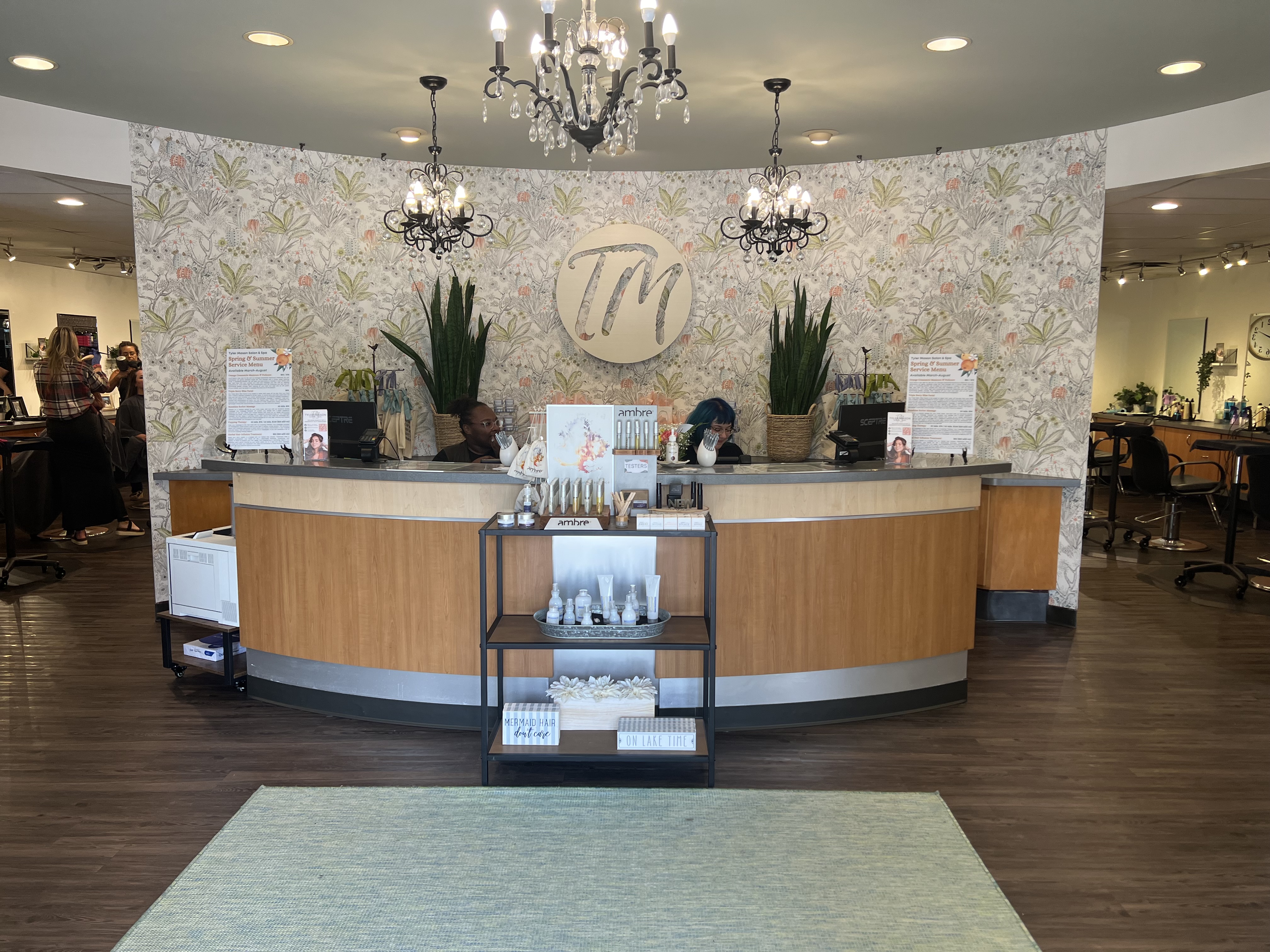 Reception area of a salon with two staff members behind a curved wooden desk, decorative plants, products on display, and a sign with the initials "TM" on the wall. - Tyler Mason Salon + Spa in Indianapolis, IN