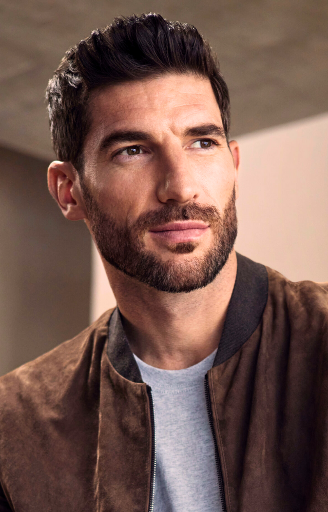 A man with short dark hair and a beard wears a brown jacket over a light gray shirt, looking slightly to the side in a softly lit indoor setting. - Tyler Mason Salon + Spa in Indianapolis, IN