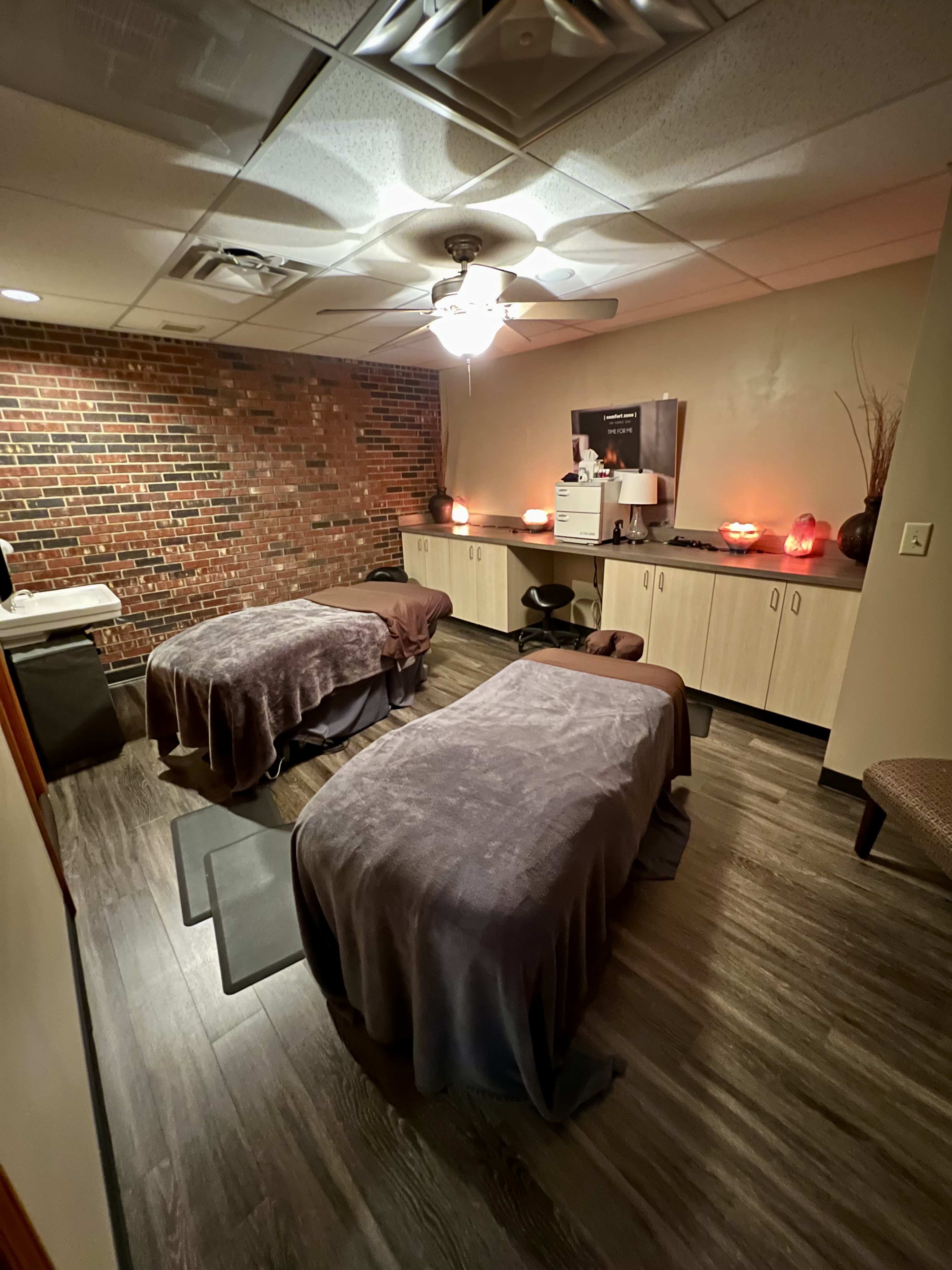 A dimly lit massage room with two massage tables covered in dark blankets, brick accent wall, cabinets, and salt lamps creating a relaxing atmosphere. - Tyler Mason Salon + Spa in Indianapolis, IN