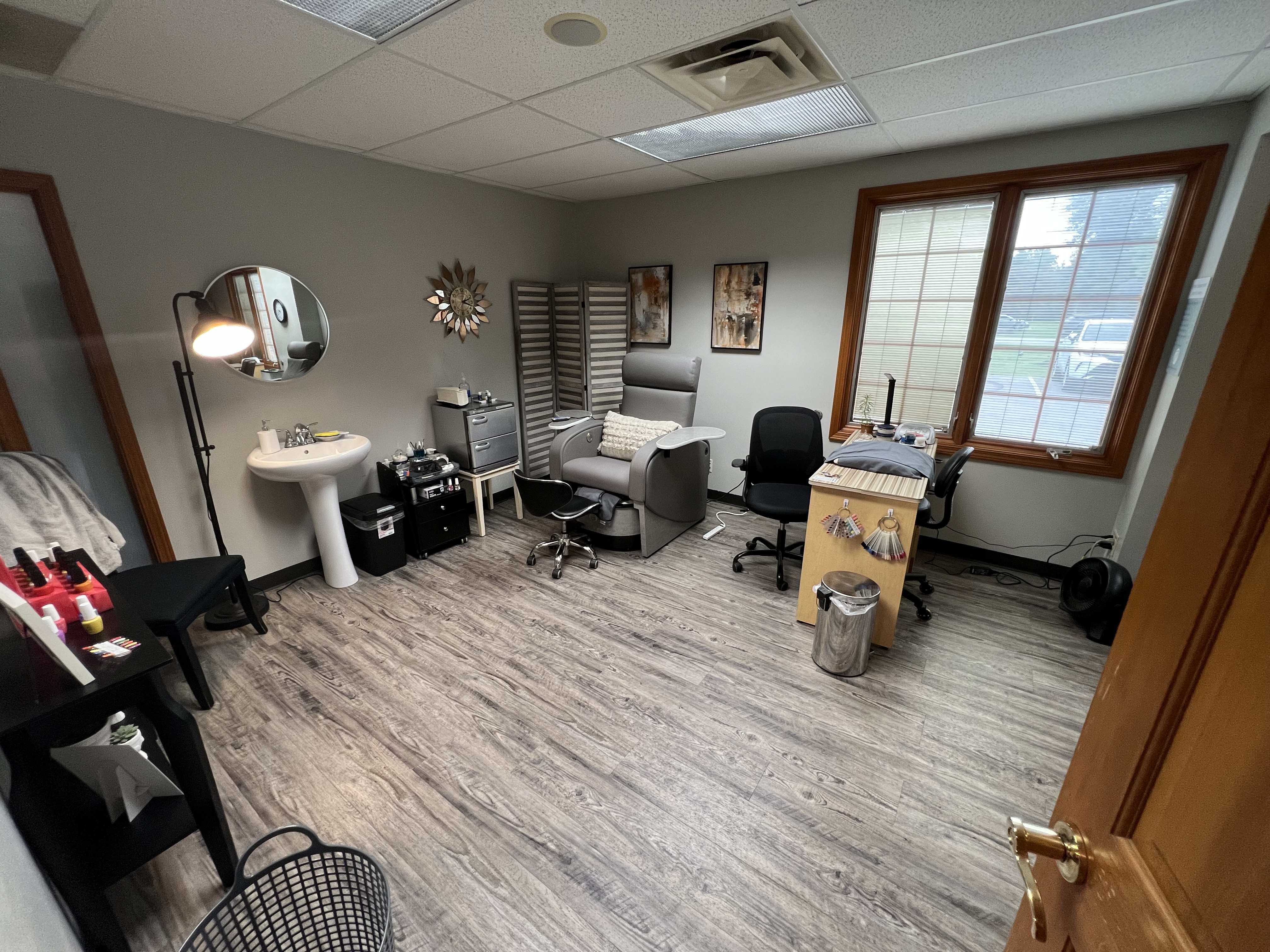A tidy salon room with a pedicure chair, manicure table, sink, mirror, and various beauty supplies, featuring wood floors and large windows letting in natural light. - Tyler Mason Salon + Spa in Indianapolis, IN