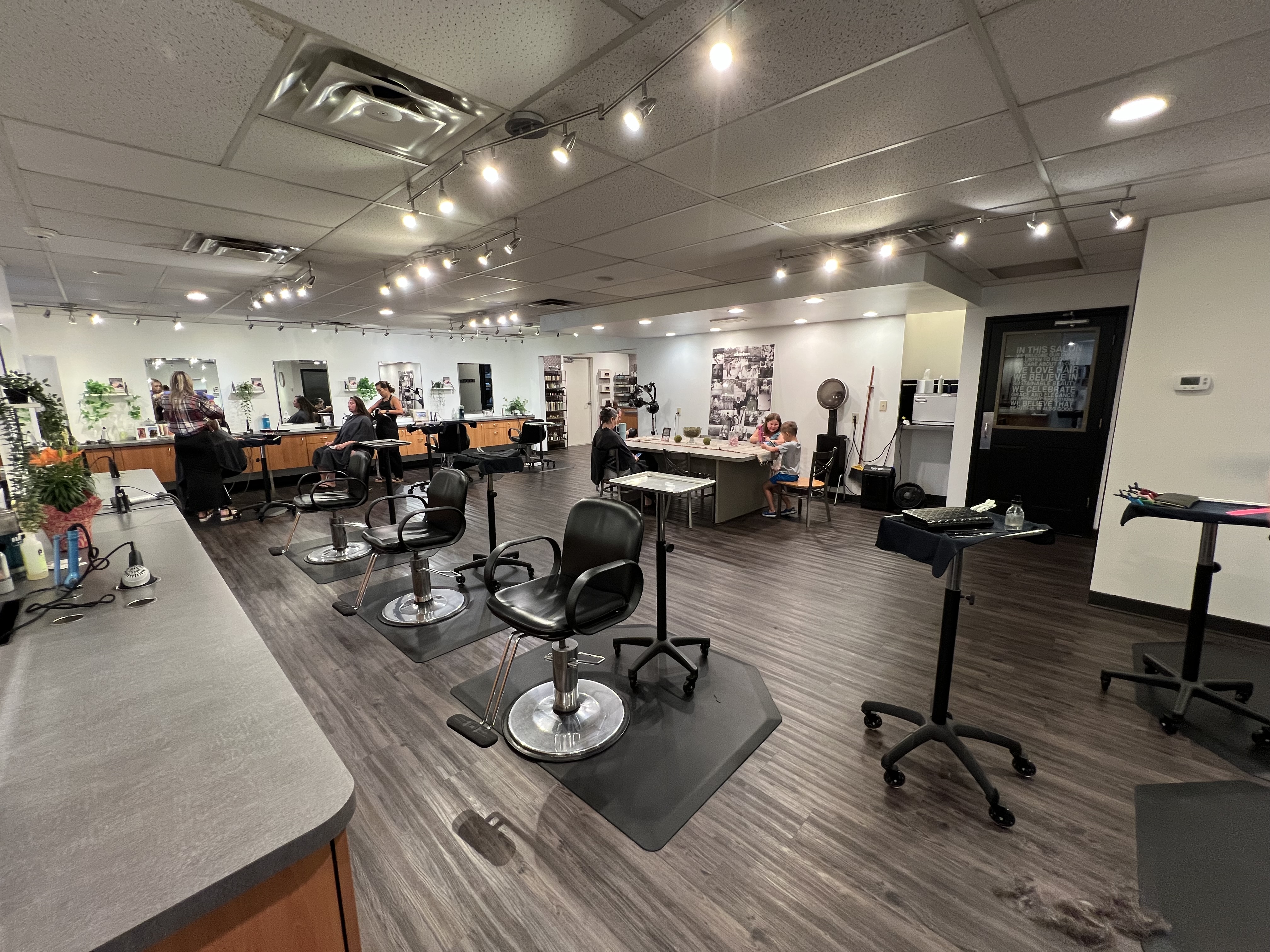 A modern salon with empty black styling chairs, a few people seated at workstations, and bright overhead lighting. The space has wood flooring and large mirrors along the walls. - Tyler Mason Salon + Spa in Indianapolis, IN