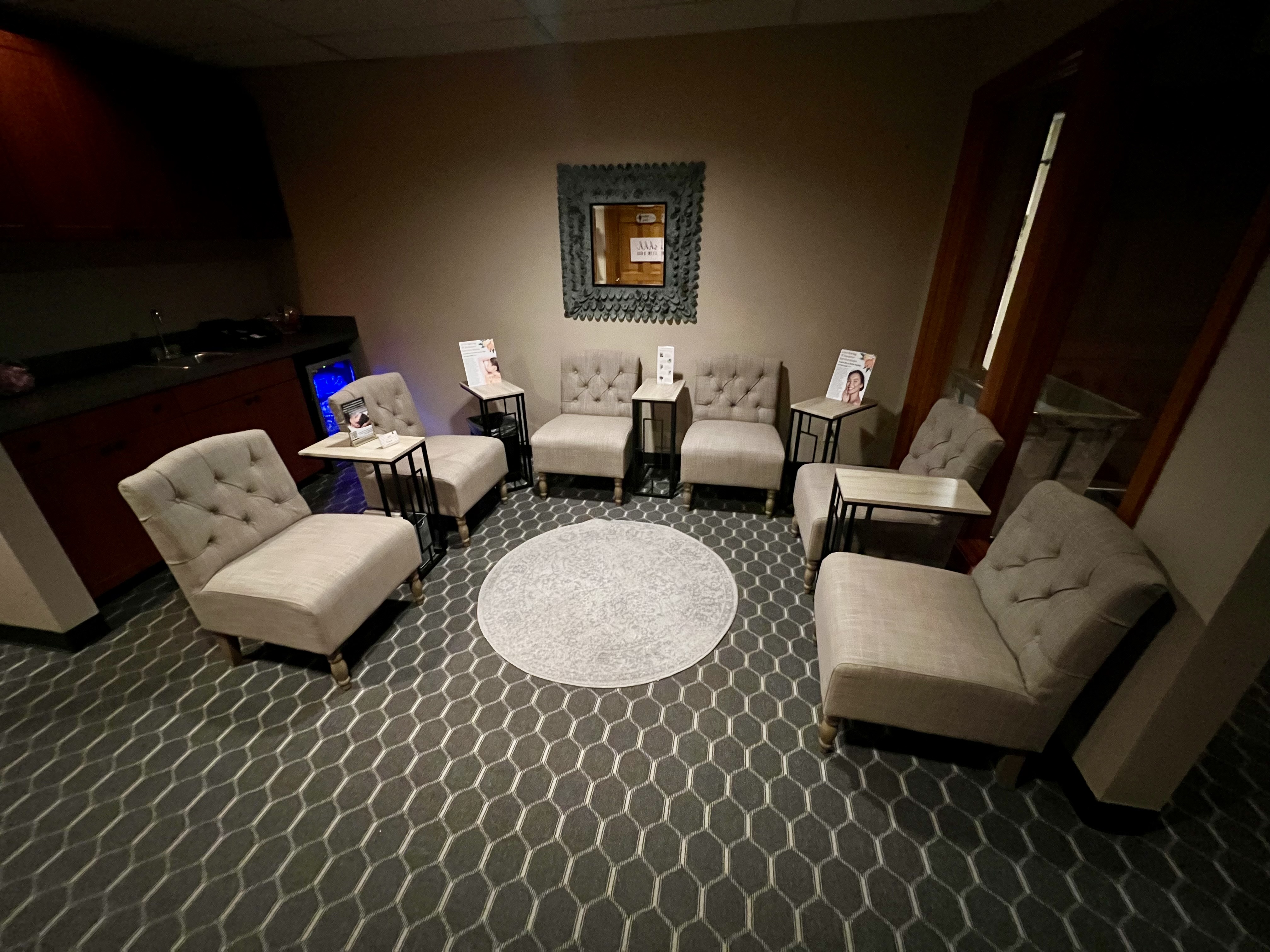 A waiting room with seven beige chairs arranged in a semicircle around a round rug, with side tables and magazines on some chairs, and a mirror on the wall. - Tyler Mason Salon + Spa in Indianapolis, IN