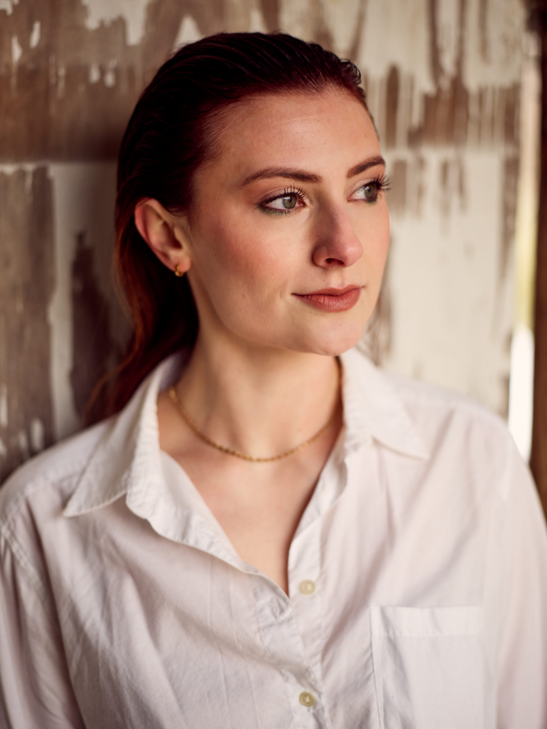 A woman with auburn hair wearing a white shirt and gold necklace stands indoors against a weathered, textured wall, looking to the side. - Tyler Mason Salon + Spa in Indianapolis, IN